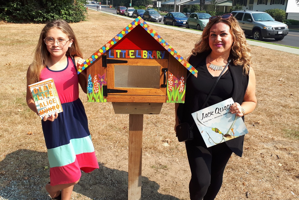 A mother and daughter stand in front of a little library.