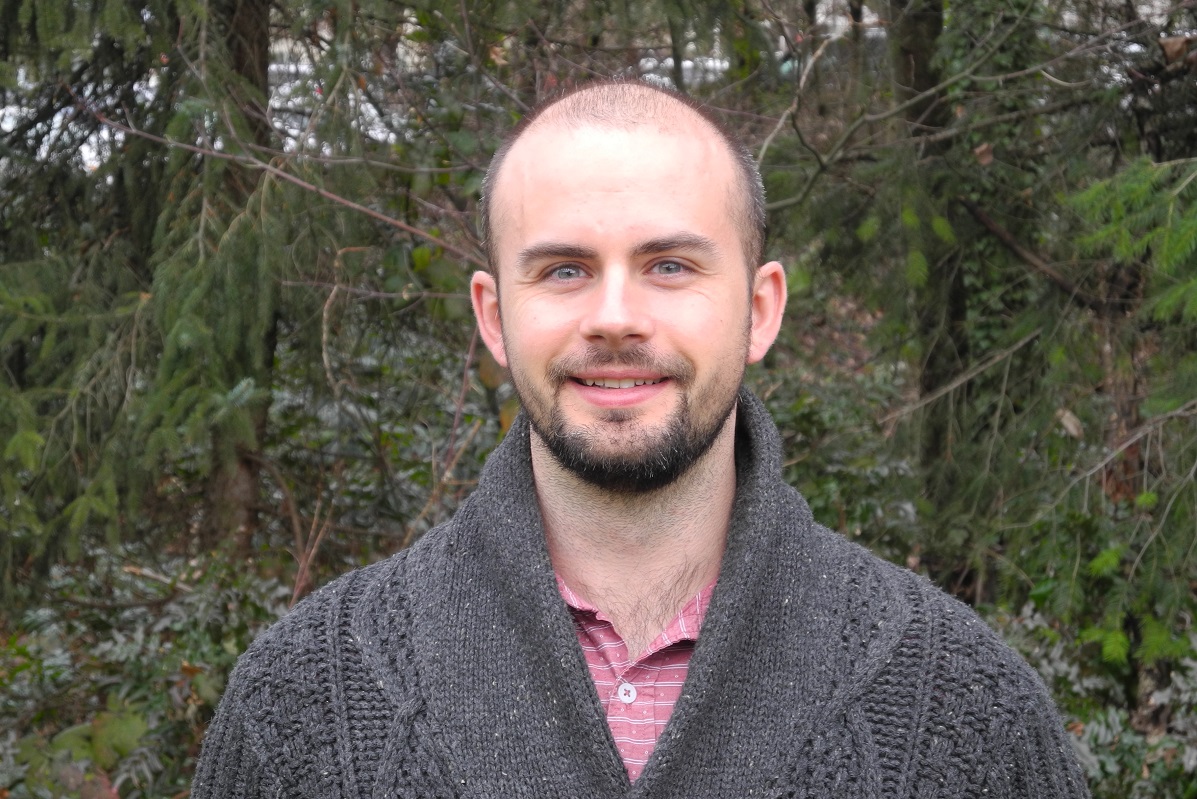 A man in a winter sweater stands in front of a forest, smiling at the camera.