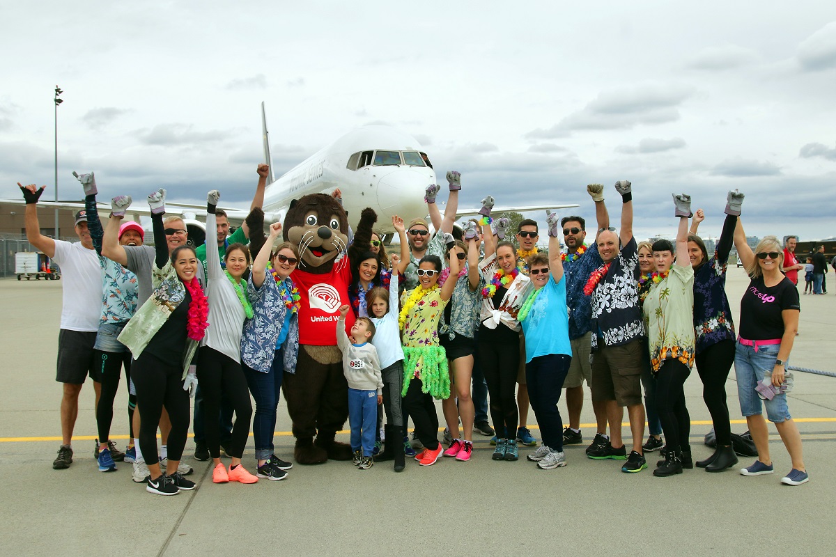 A group of exciting, smiling people wear Hawaiian shirts and pose on a tarmac in front of a plane.