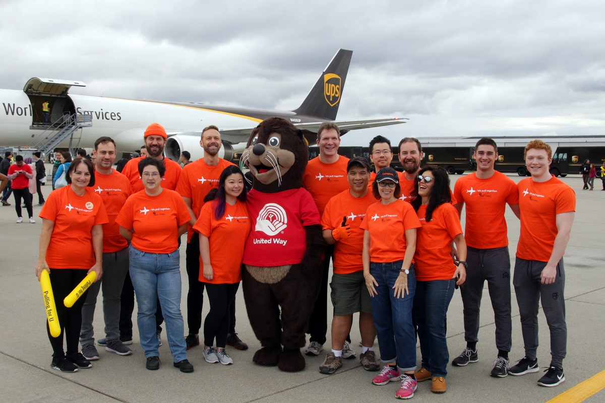 A smiling group of individuals all wear orange t-shirts and stand in front of a plane.