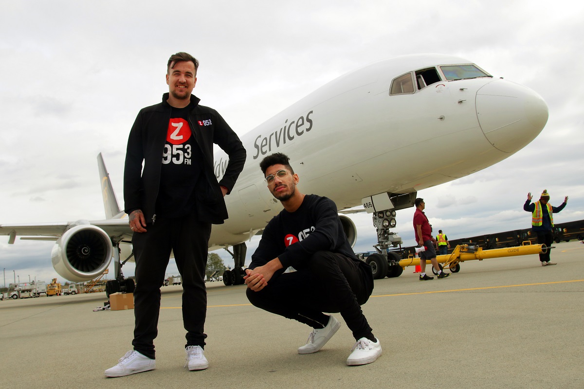 Two men in matching outfits pose on a tarmac in front of a plane.