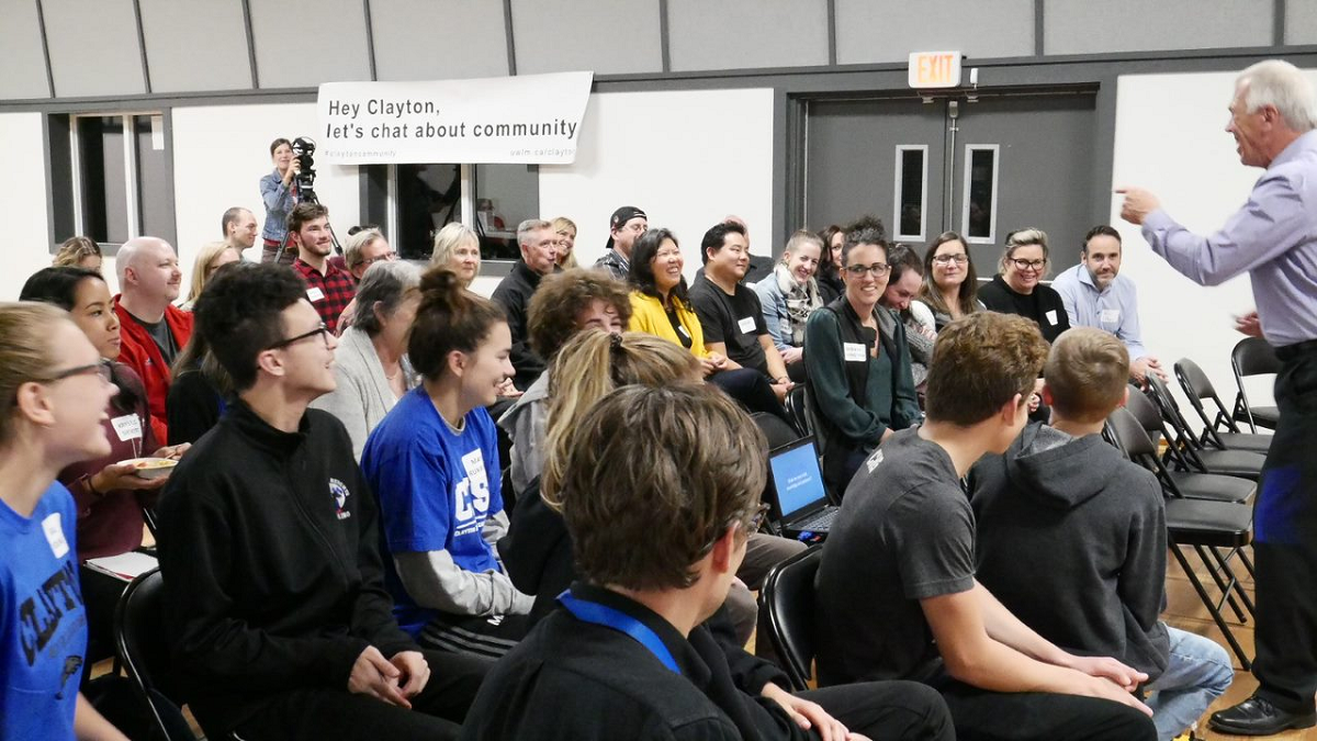 A man stands at the front of a room and speaks to a crowd of people.