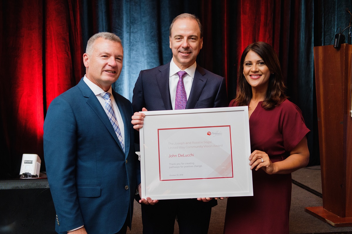 Two men in suits stand next to a woman. One of the men holds a framed award.
