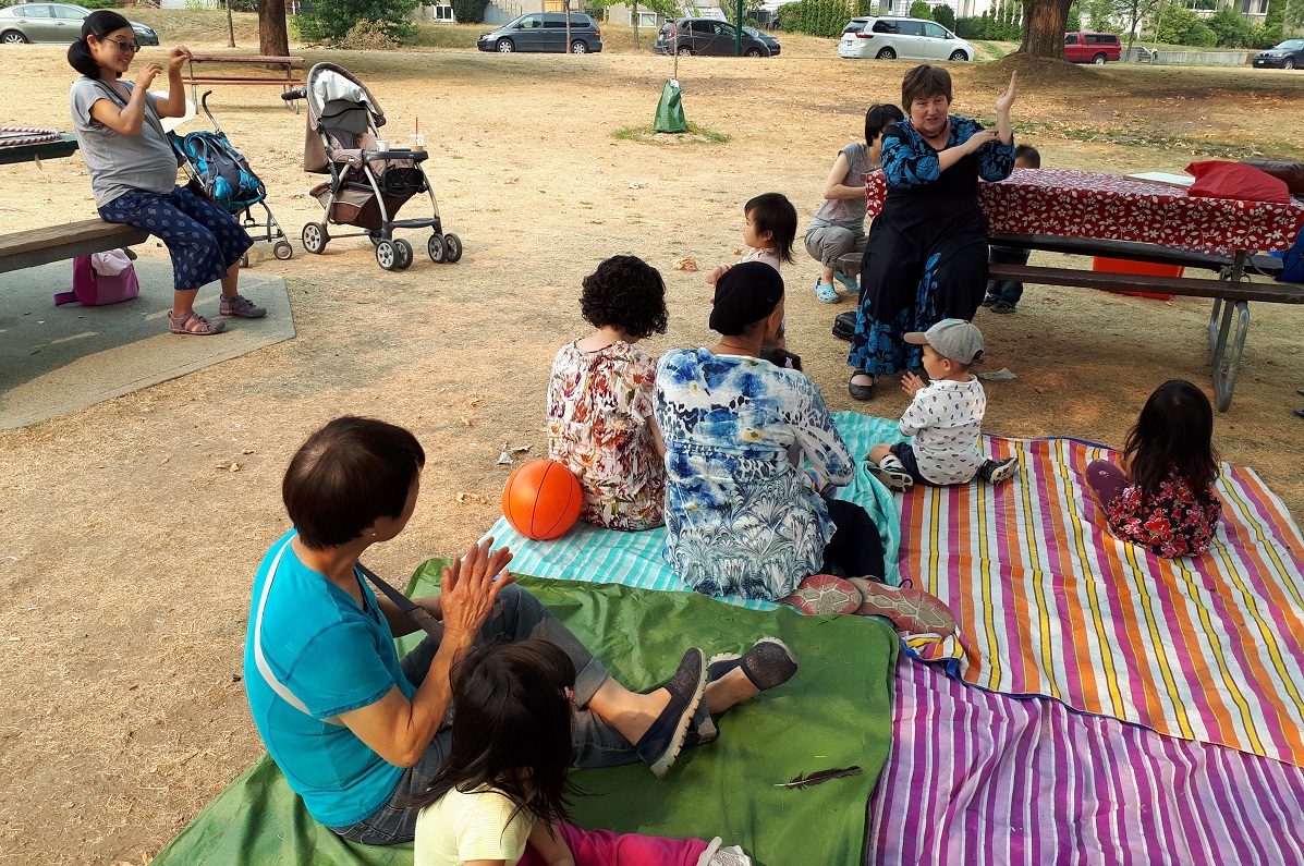 A group of parents and caretakers sit with children listening to a story in a park.