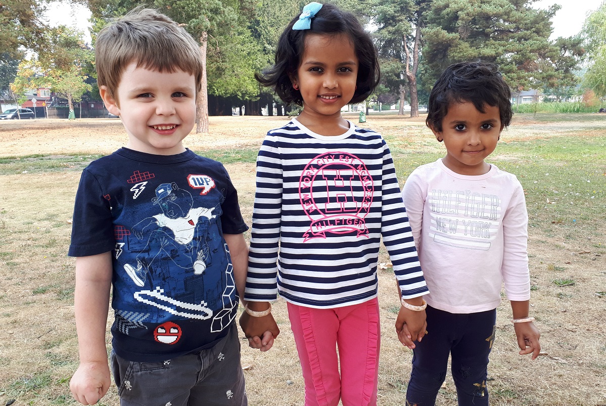 Three children, a boy and two girls, hold hands and smile at the camera.