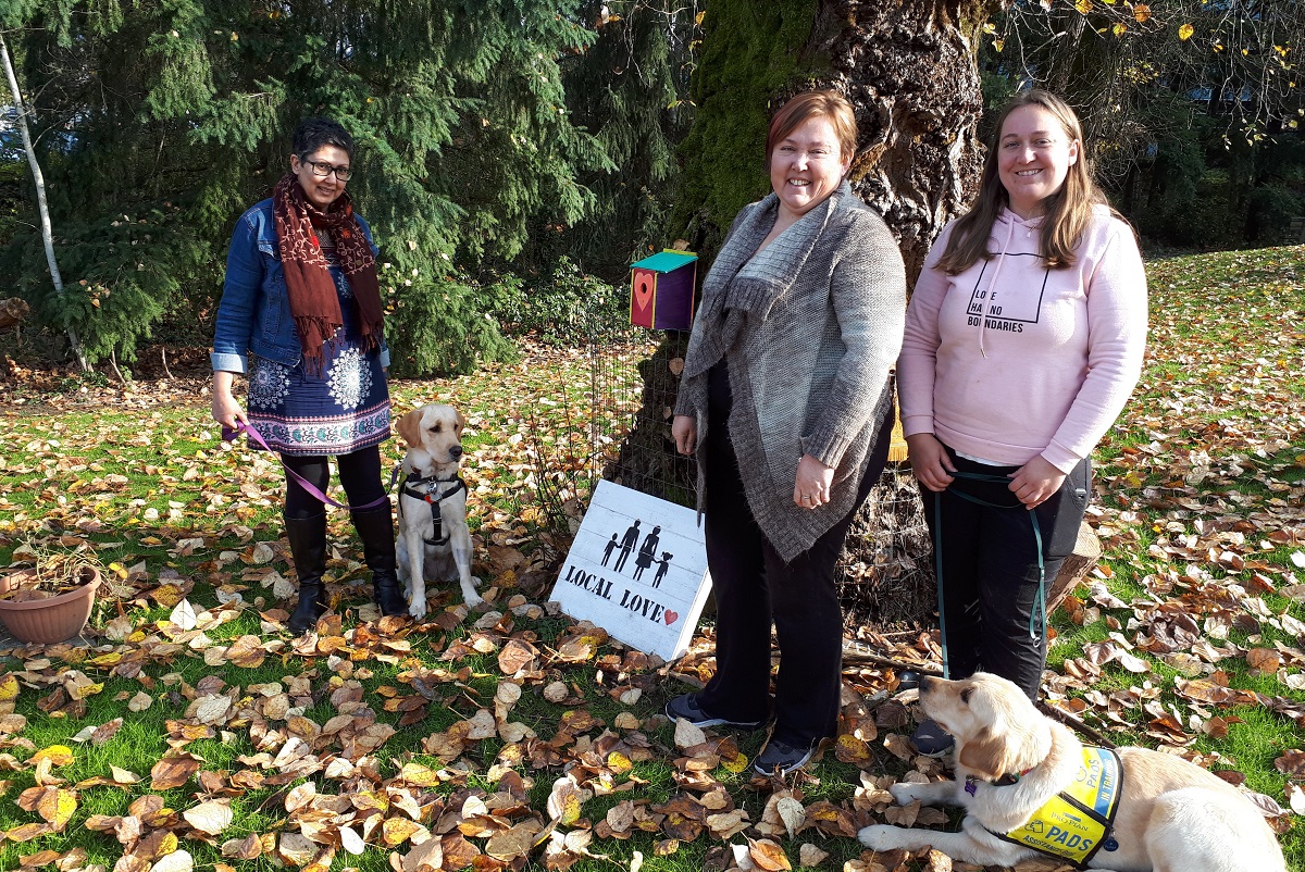 Three women stands outside with two golden retrievers.