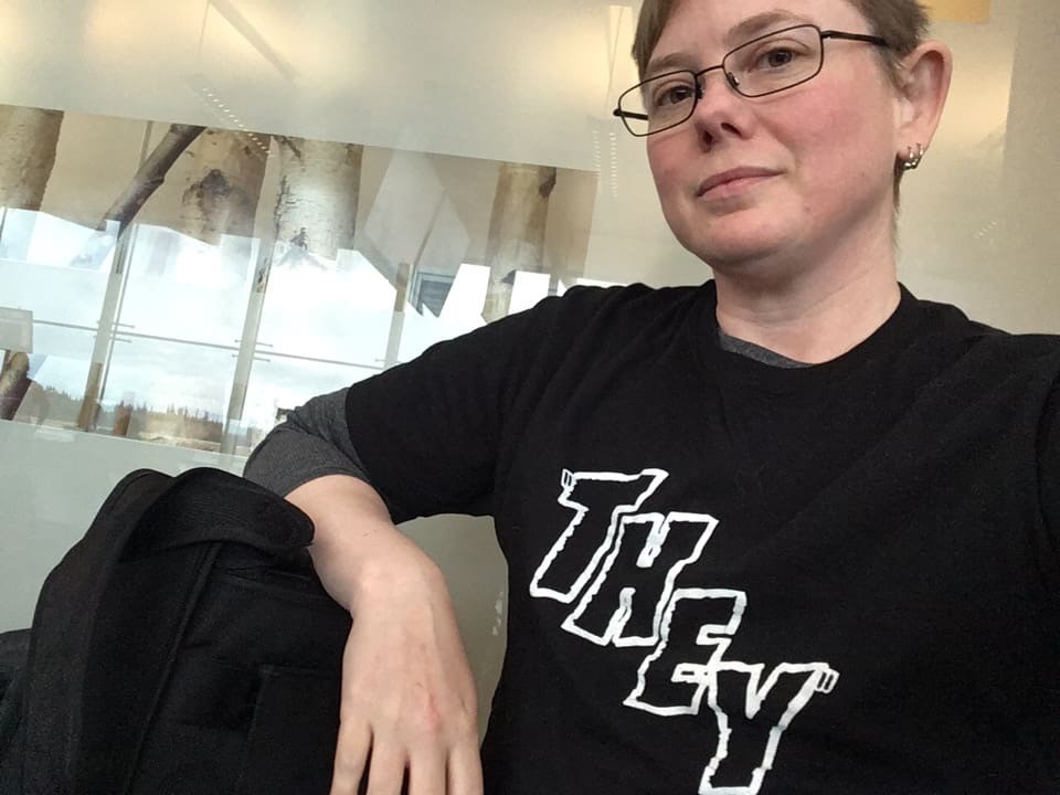 A person wears a black sweater with the word "they" on the front.