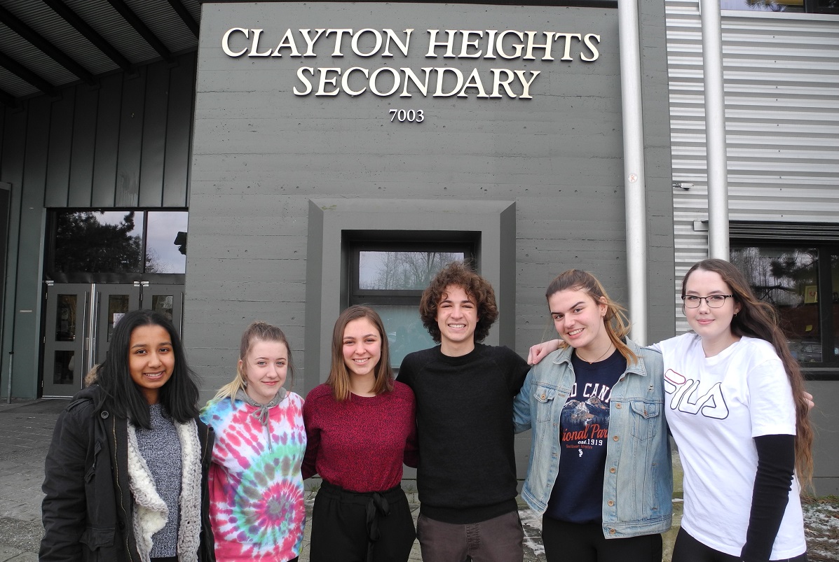 A group of teenagers stand outside of a high school and smile at the camera.